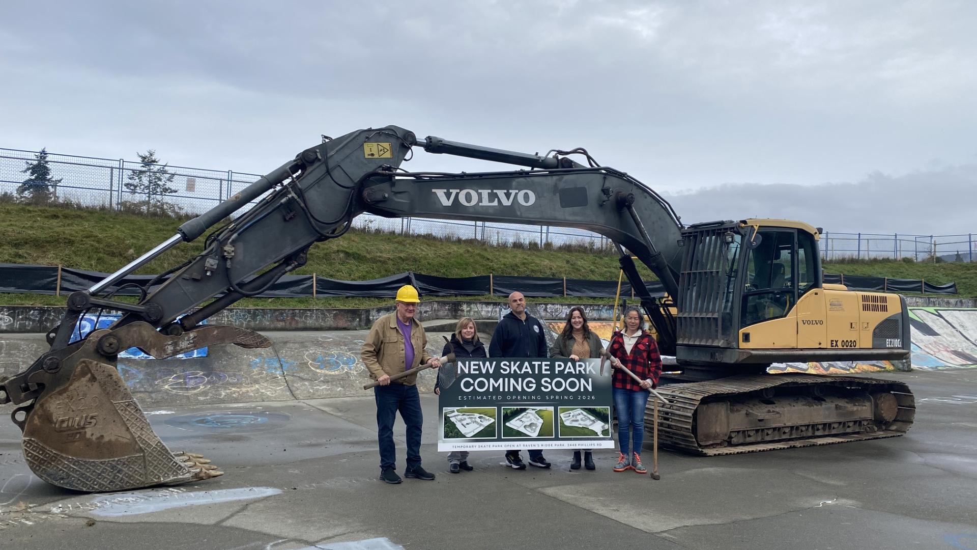 Three commission members and two staff standing in front of an excavator in the skate park.
